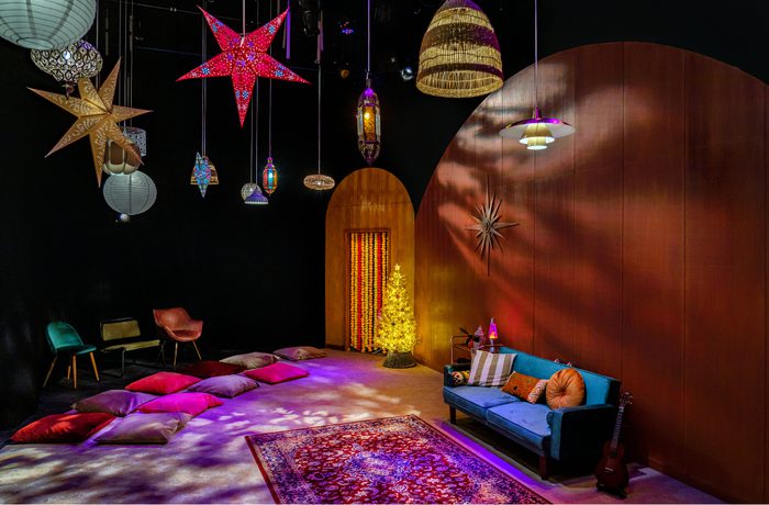 A cozy room with a festive vibe, featuring colorful star lanterns, a glowing tree, cushions on the floor, a blue sofa, and a guitar against warm wood walls. This photo is of the set from cast's 2021 production of Wish, a early years production in Drama Space.