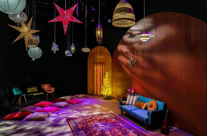 A cozy room with a festive vibe, featuring colorful star lanterns, a glowing tree, cushions on the floor, a blue sofa, and a guitar against warm wood walls. This photo is of the set from cast's 2021 production of Wish, a early years production in Drama Space.