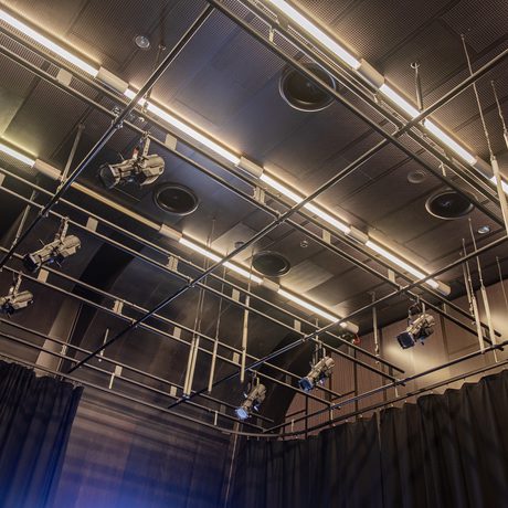 Ceiling view of a Drama Space at Cast, featuring pendulum house lights, a grid of stage lights and equipment suspended from metal beams, with a backdrop of black curtains.