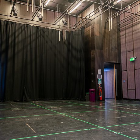 Large, empty Drama Space with black curtains, green-taped floor grid, and a doorway leading to a well-lit hallway. Calm, professional atmosphere.