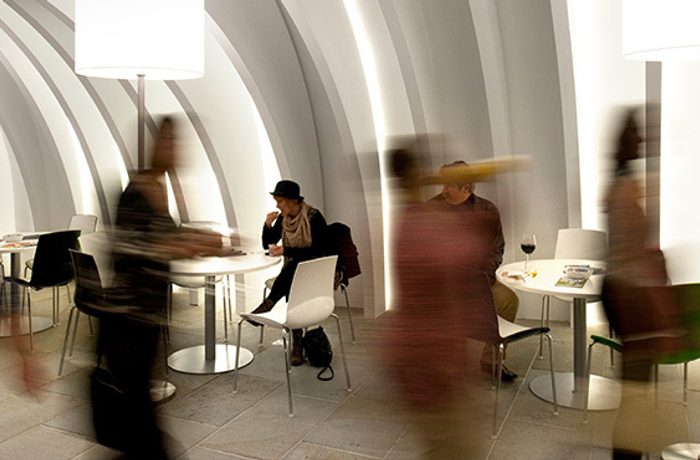 A stylistic blurry shot of people moving through the Cast foyer. A woman in focus in the background is wearing a black bowler style hat, resting her elbow on a table.