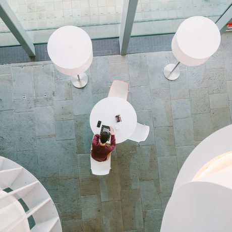 A shot taken from above, looking down on two standing lights and a single person sat at a table reading. The image is clean and bright.