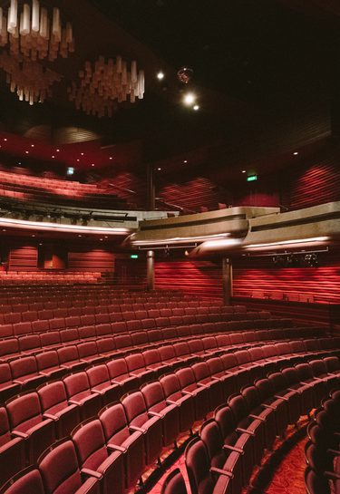A spacious, empty theater auditorium in Cast's Main Space with curved rows of plush red seats, warm lighting, creating a cozy, inviting ambiance.