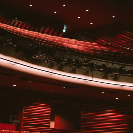 Dimly lit theater circle in Cast's Main Space with rows of red seats, overhead lighting equipment, and an exit sign glowing green. The atmosphere is warm and serene.