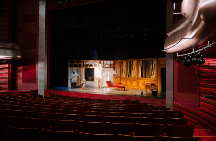 Cast's Main Space stage with an elegant, vintage set featuring a red chaise lounge, table, and backdrop with curtains. Empty red seats create a quiet, anticipatory mood.