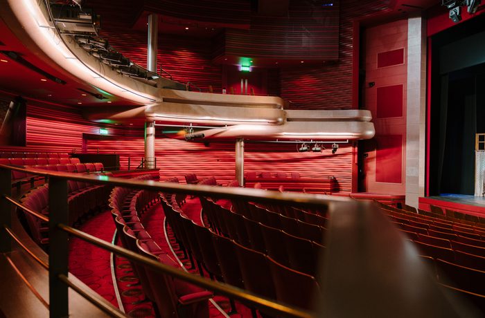 Empty auditorium in Cast's Main Space with rows of red seats, curving balcony, and stage on the right. Warm lighting creates a cozy, dramatic atmosphere.