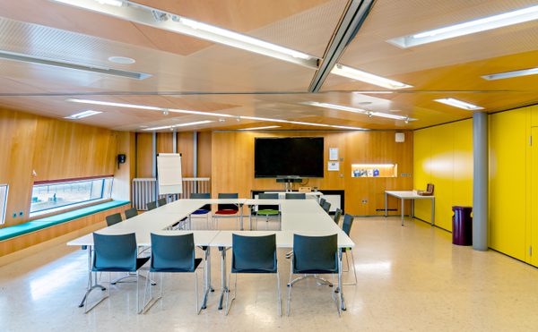 Cast's modern Meeting Space with white tables in a rectangular formation, black chairs, wooden walls, and a large screen. Bright yellow accents and ambient lighting.