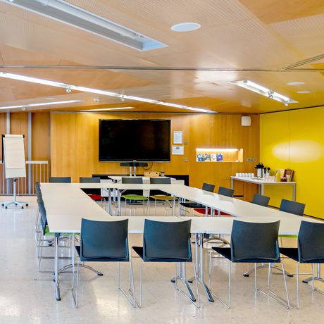 Cast's modern Meeting Space with a rectangle-shaped table arrangement, black chairs, a large wall-mounted TV, flip chart, and refreshments on a side table.