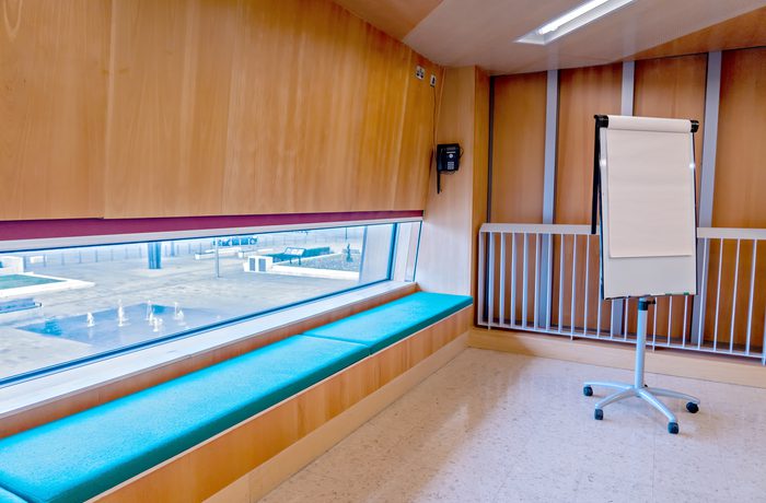 Corner of Cast's Meeting Space, with a teal cushioned window seat, and a blank flip chart on a stand. Large window offers a view outside of an empty Sir Nigel Gresley square and fountain.