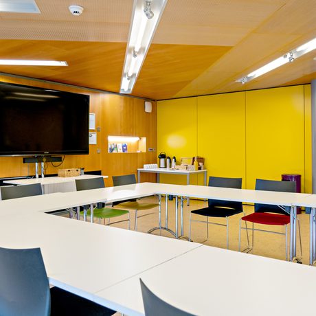 Cast's modern conference room Meeting Space, with rectangular-shaped white tables and various colored chairs. A large black TV is mounted on the wood-paneled wall. Bright yellow accents add vibrancy.