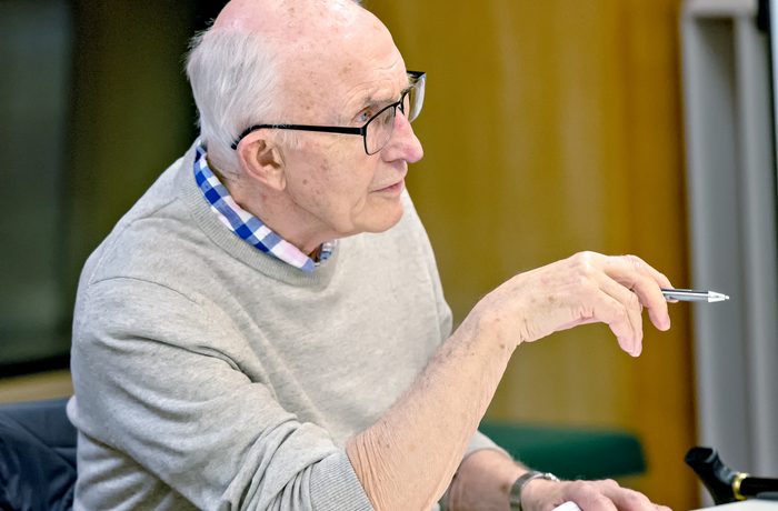 Older man in glasses, gray sweater, and checked shirt ponders while holding a pen and notepad in a scriptworks session in Cast's Meeting Space, conveying focus and contemplation.