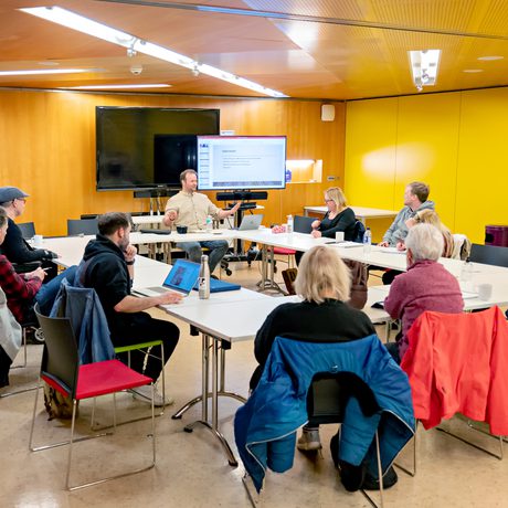 A diverse group of people sitting around tables in Cast's meeting room with wooden walls and yellow accents. A presenter leads the discussion, gesturing towards a screen during a session of Scriptworks..