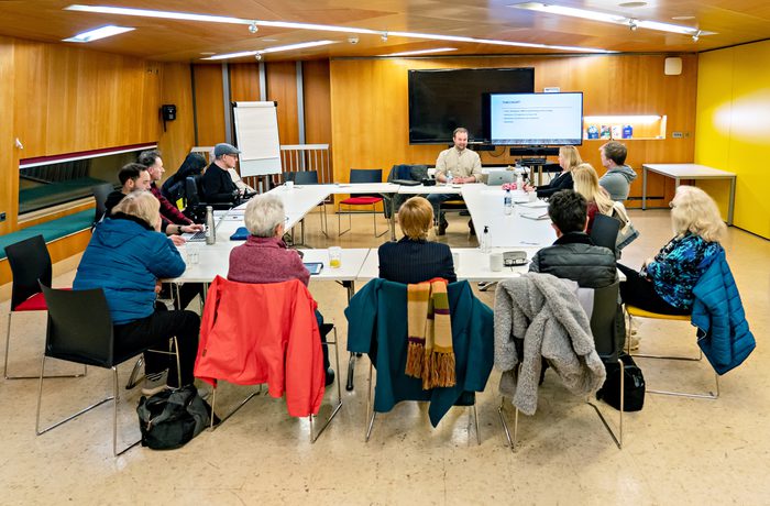 A group of people sit in a U-shaped formation around tables in a conference room, attentively listening to a speaker near a large screen displaying a presentation. Photo taken during a Scriptworks session at Cast in their Meeting Space.