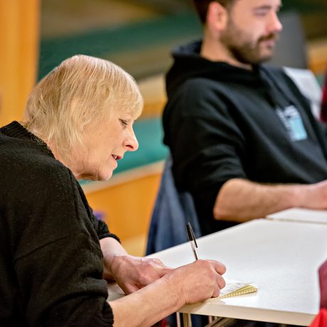 A woman with short blonde hair attentively writes in a notebook at a table, while a man in the background listens, creating a focused atmosphere in Cast's Meeting Space.