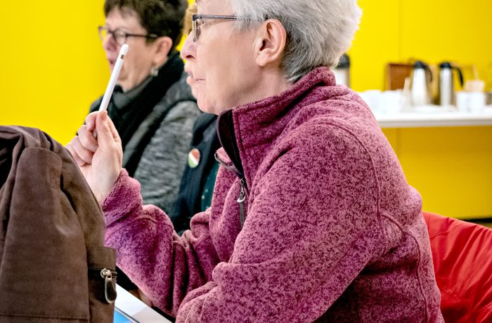 A person with short white hair and glasses, wearing a pink sweater, holds a pen thoughtfully in a bright room with yellow walls and a drink on the table during a session of Scriptworks at Cast in Meeting Space.