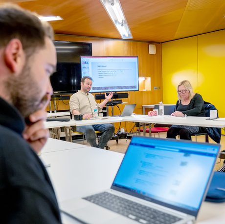 A man presents to a focused group in a meeting room with yellow walls. Attendees take notes, and one uses a laptop, creating a collaborative atmosphere during a session of Scriptworks at Cast in their Meeting Space.
