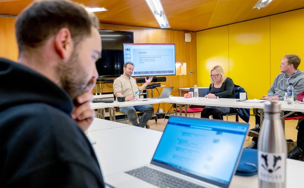 A man presents to a focused group in a meeting room with yellow walls. Attendees take notes, and one uses a laptop, creating a collaborative atmosphere during a session of Scriptworks at Cast in their Meeting Space.