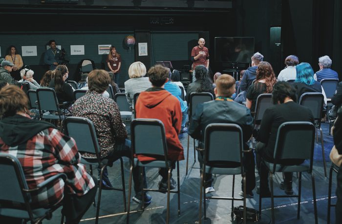 Audience seated in a dimly lit room listens to a speaker near a screen with "Open" and "Brave" signs along the wall. Engaged and focused atmosphere during the Public Acts Celebration Day in Cast's Second Space. .
