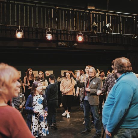 A diverse group of people, including children and adults, gather in a dimly lit theater, smiling and interacting energetically, creating a lively, joyful atmosphere during Public Acts Celebration Day held in the Second Space at Cast.