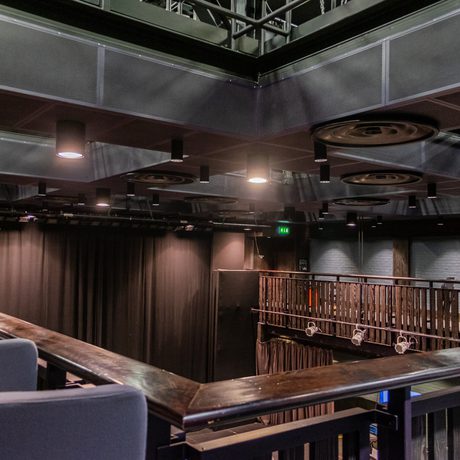 Dimly lit theatre balcony with gray seats and a view of a stage curtain inside Cast's Second Space, a box studio theatre. Overhead, circular spotlights create an industrial atmosphere.