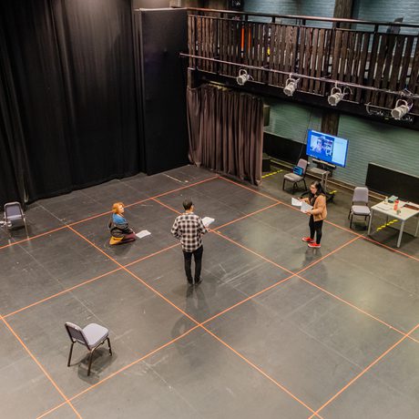Aerial view of a rehearsal in a black box theatre setting. Three people spaced apart during Covid-19 restrictions on a taped floor grid, holding scripts. A screen and empty chairs are visible. The photo was taken in Cast's Second Space.