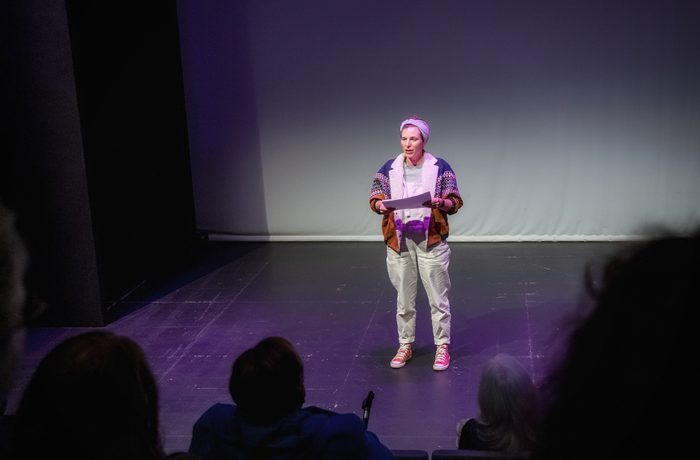 A person stands on a dimly lit stage holding papers, facing an audience. The setting is minimalist, conveying a serious, focused tone during Cast's event called Fresh Works Scratch Night held in the Second Space.