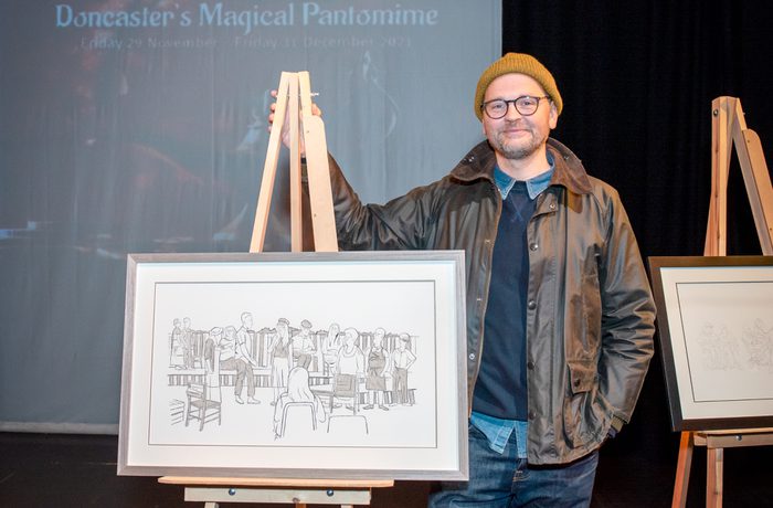 A person wearing a yellow beanie and glasses stands beside an easel displaying a line drawing of people during Cast's exhibition in the Second Space called Here and Now. Background text reads "Doncaster's Magical Pantomime."