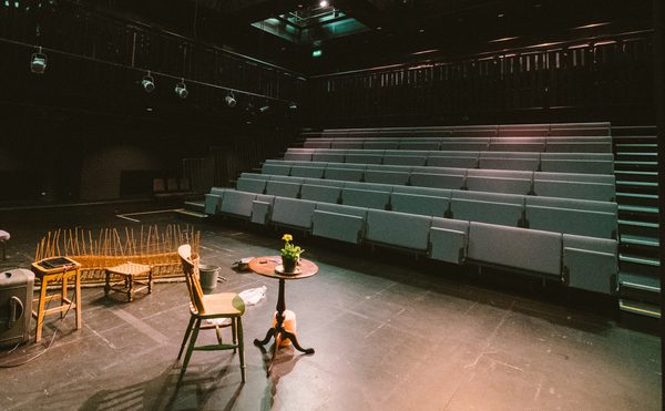 An empty stage with dim lighting and modern seating inside Cast's Second Space. A few chairs, a small table with a plant, and wooden props set a calm, anticipatory mood.
