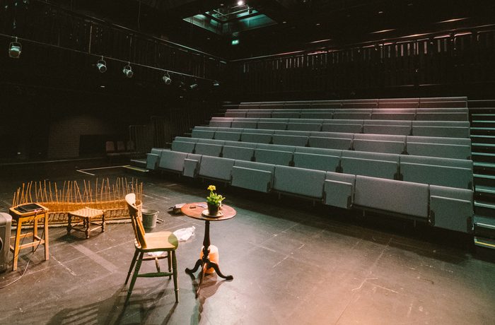 An empty stage with dim lighting and modern seating inside Cast's Second Space. A few chairs, a small table with a plant, and wooden props set a calm, anticipatory mood.