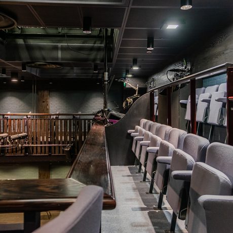 Dimly lit auditorium in Cast's Second Space. The image shows tiered, balcony seating with gray cushions and wooden railings. Spotlights angled towards a stage area convey a sense of anticipation