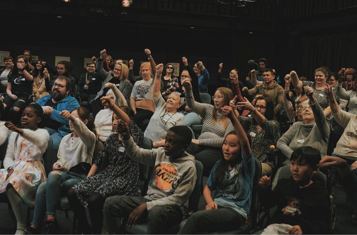 A diverse group of people in an auditorium, smiling and raising their fists enthusiastically. The atmosphere is joyful and united, conveying a sense of community during the Public Acts Celebration Day at Cast in the Second Space.
