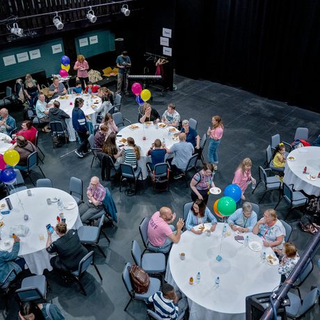 Aerial view of a lively gathering with people seated at round tables covered in white cloths. Colorful balloons and a celebratory atmosphere fill the room during the Public Acts Celebration Day held in Cast's Second Space.